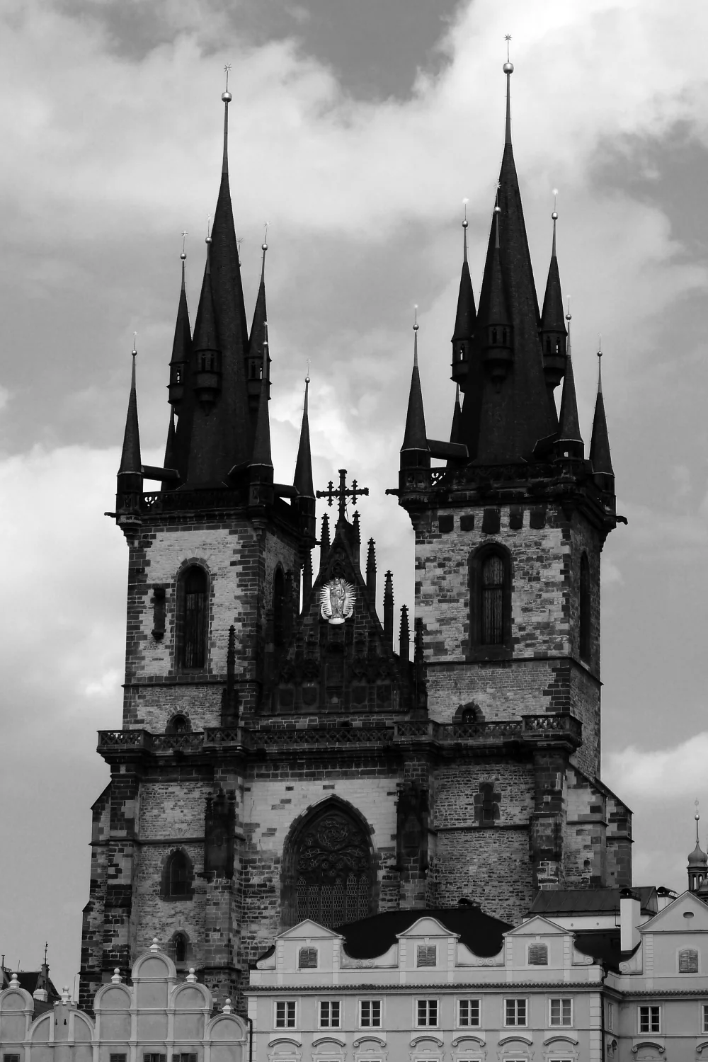 Týn Church in Prague, in black and white: its two Gothic towers, each crowned with a cluster of dark spires, rise against a cloudy sky above the gabled houses of Old Town Square.